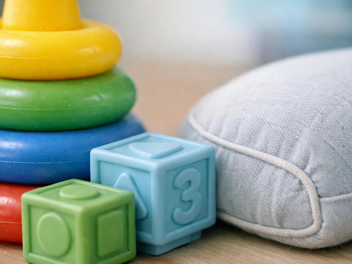 Colorful baby toys, stacking rings and number blocks beside a gray cushion on a wooden floor