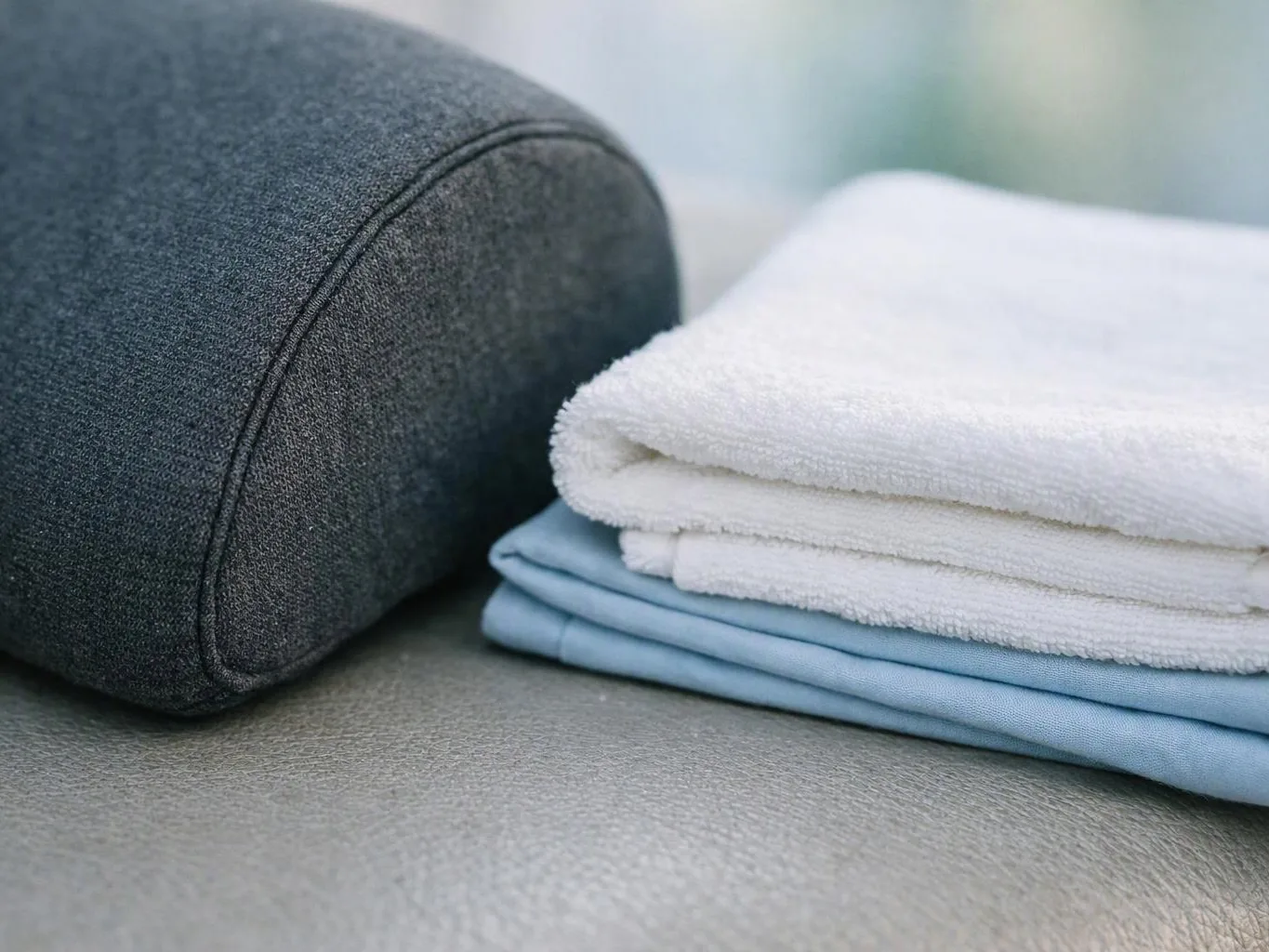 A folded stack of white and blue towels sits beside a dark gray fabric pillow on a couch