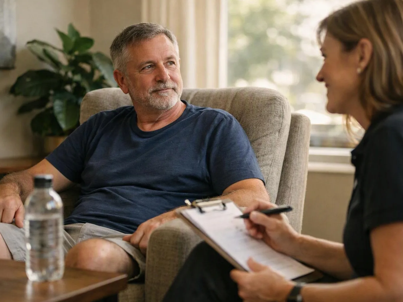 A man sits in a chair talking with a woman holding a clipboard in a bright living room
