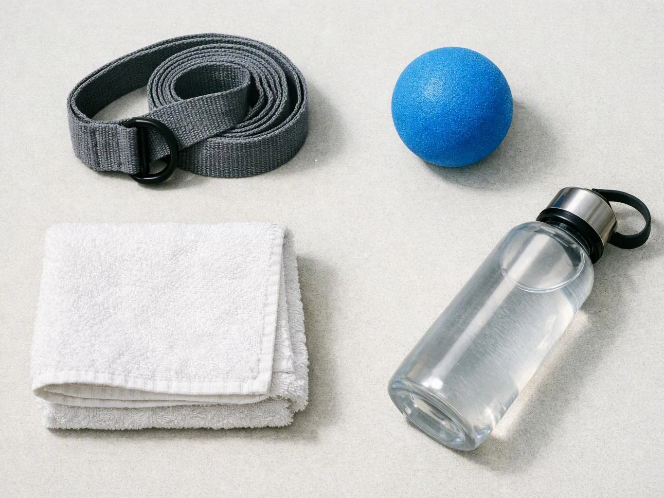 A rolled gray strap, blue massage ball, folded white towel, and clear water bottle sit on a light surface